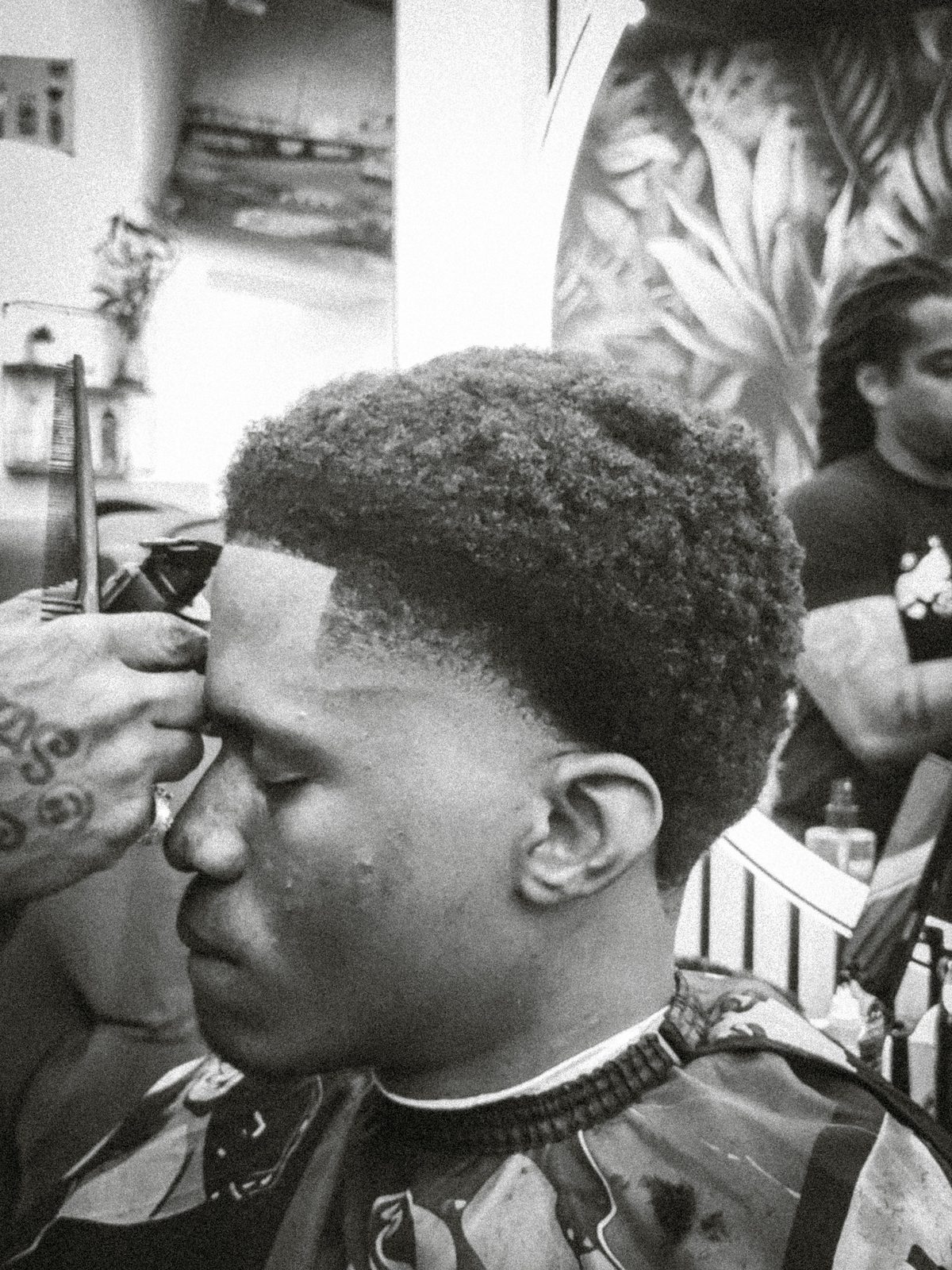 Black-and-white close-up of a young man mid-haircut, eyes closed, as a barber's tattooed hand lines up his hairline with clippers.