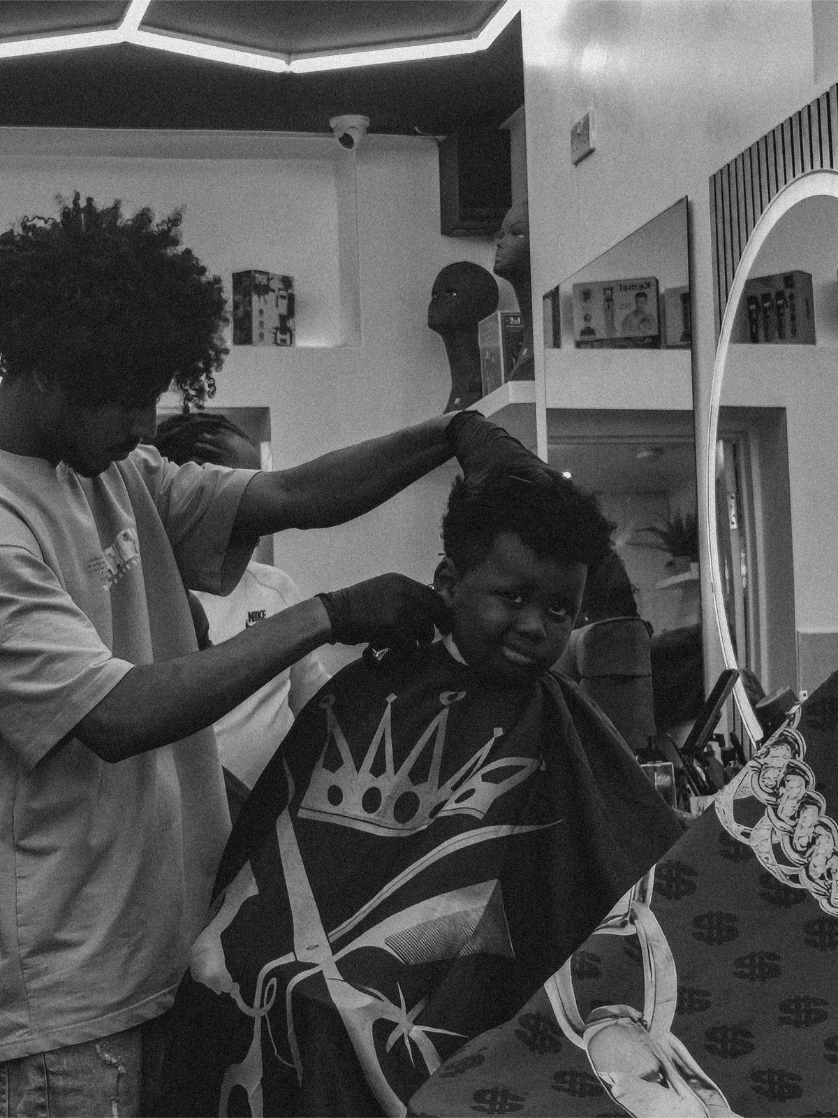Black-and-white photo inside a barbershop: a barber cuts a young boy's hair while the boy sits under a patterned cape.