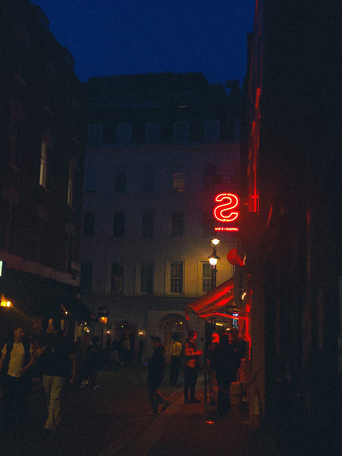 A narrow street at night, people gathered outside lit bars, a red neon sign glowing on the right.