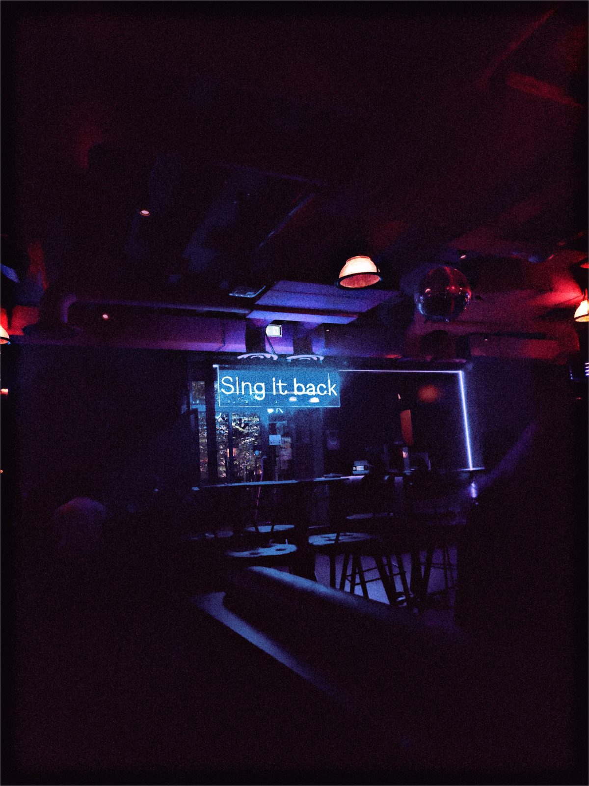 A dim bar in purple and red light, a blue neon sign reading &ldquo;Sing it back&rdquo; above the counter.