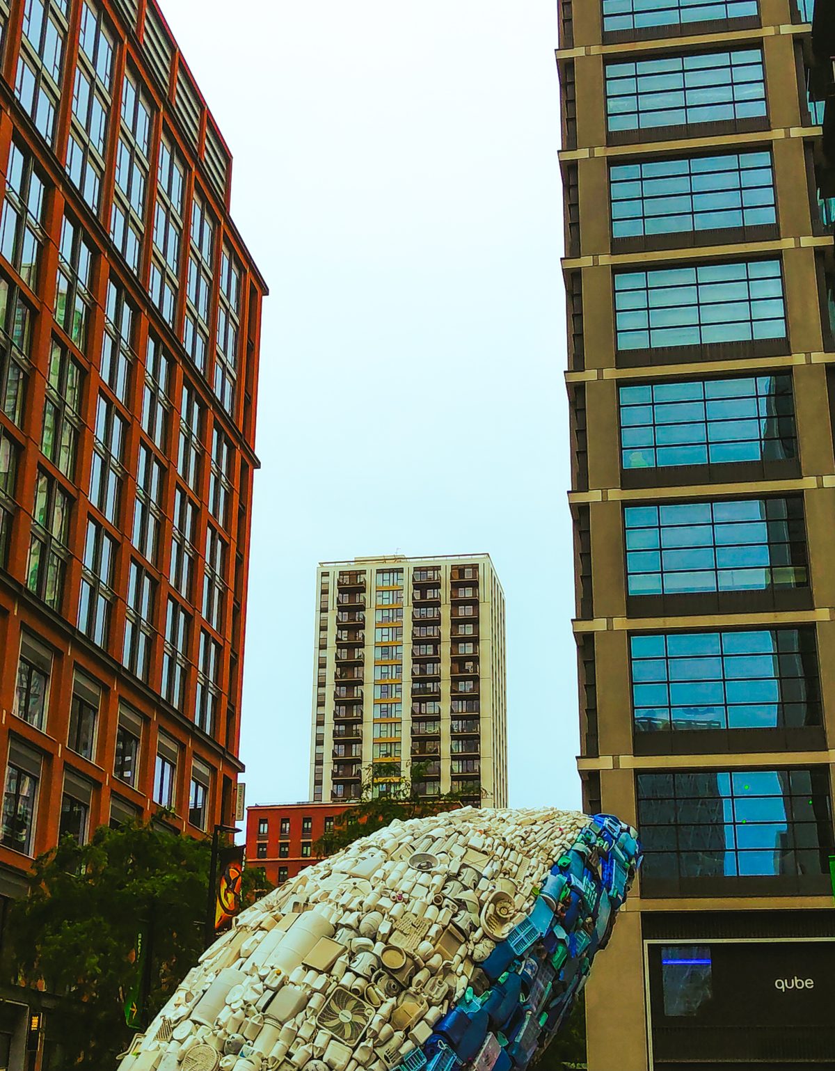 A huge sculpture clad in stone-like tiles sits between two modern office blocks, a residential tower beyond under a pale sky.