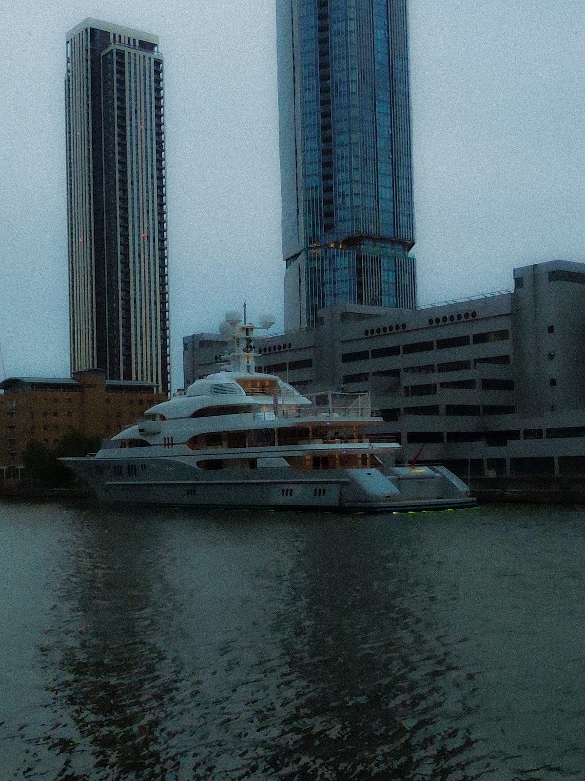 A large white yacht moored on dark water beneath two glass residential towers, under an overcast sky.