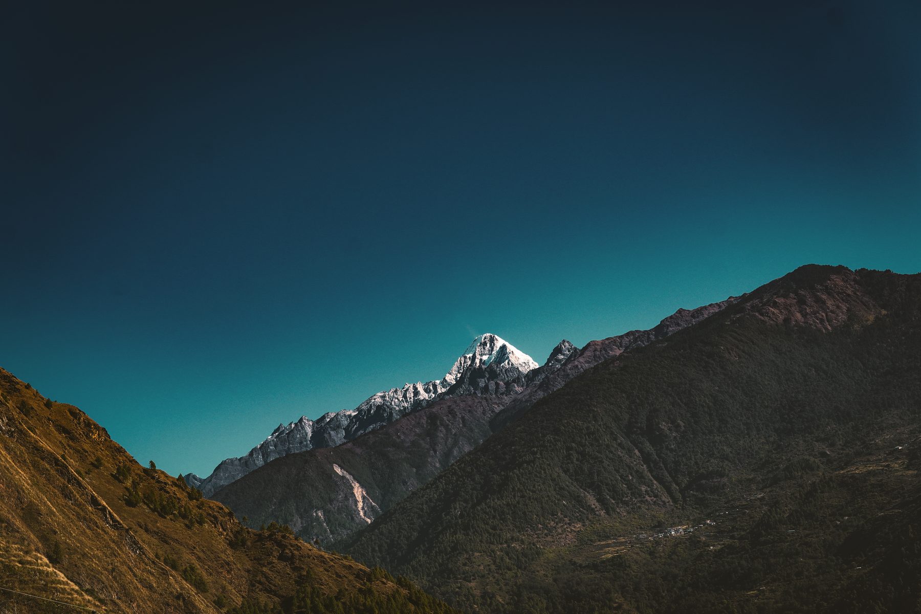 A rocky mountain ridge at dusk, a snow-capped peak catching the last light beneath a deep teal sky.
